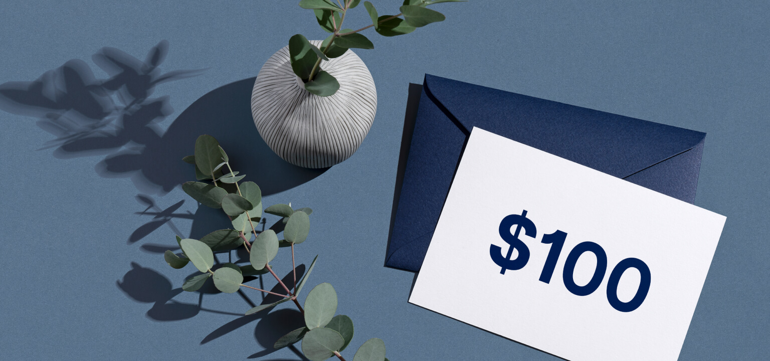 Minimalist composition with a white vase, eucalyptus branches, a navy blue envelope, and a card displaying $100