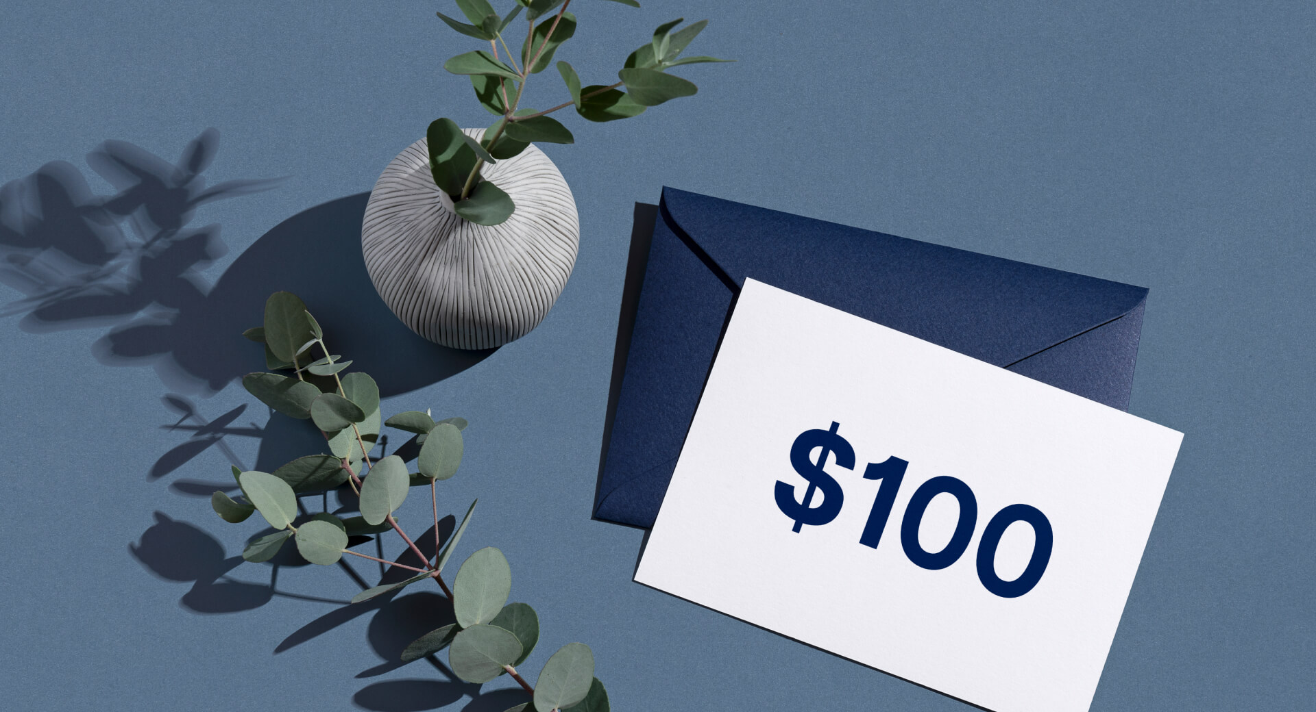 Minimalist composition with a white vase, eucalyptus branches, a navy blue envelope, and a card displaying $100